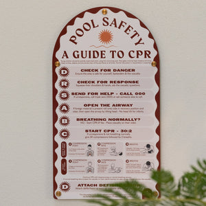 Pool safety guide to CPR on a white wall with greenery at the bottom.